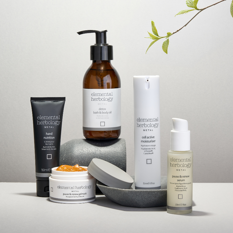 Six skincare products from elemental herbology METAL are arranged on a light gray surface with gray stones and a small leafy branch in the corner. The products include a black hand purifying tube, an amber hand body oil pump bottle, a white cell active moisturizer bottle, a beige serum bottle, and a white jar with orange cream.