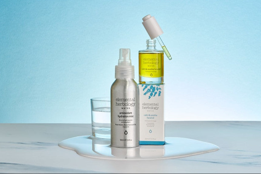 Elemental Herbology Nature hydrating hydration mask and oil set on a white marble surface with a light blue background. The set includes a silver spray bottle, a glass of water, a yellow oil bottle with a white dropper, and a white box, all branded with the 'elemental herbology nature' logo.