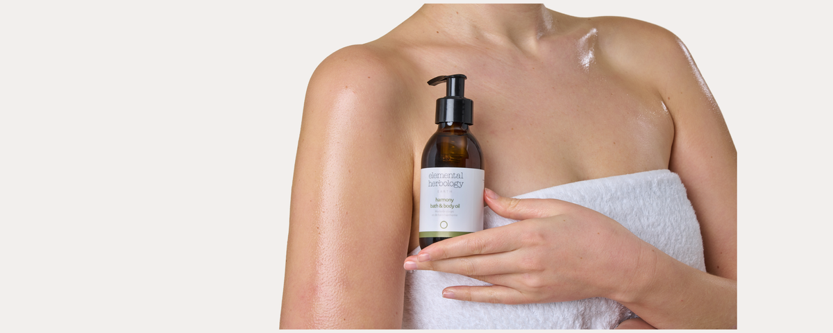A woman wrapped in a white towel holds a dark amber pump bottle with a white label against a plain light beige background.